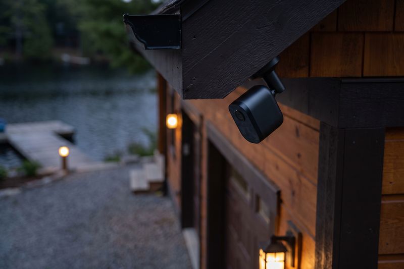 A small security camera under a cottage eave, looking down a driveway toward the dock at dusk