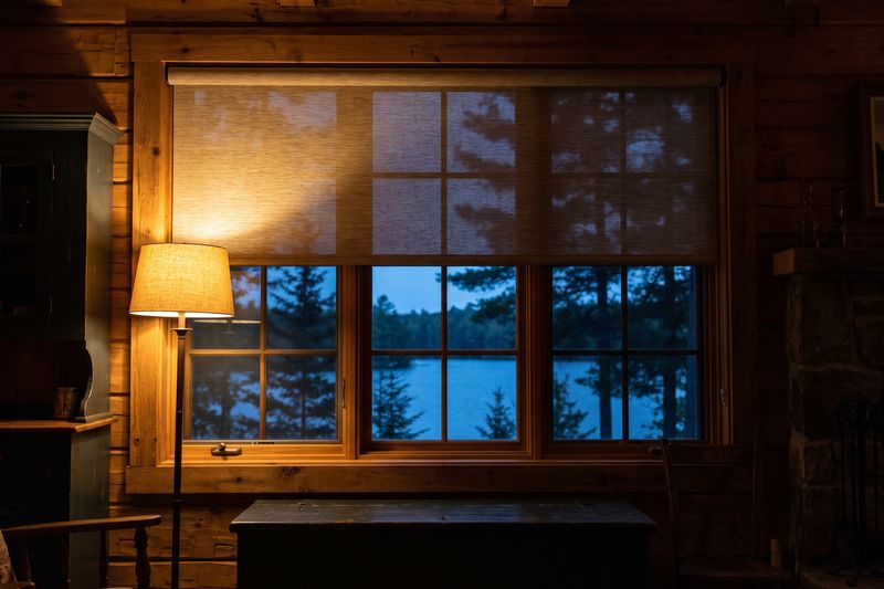 A motorised roller blind half lowered over a cottage window at dusk, warm lamp light on the timber wall